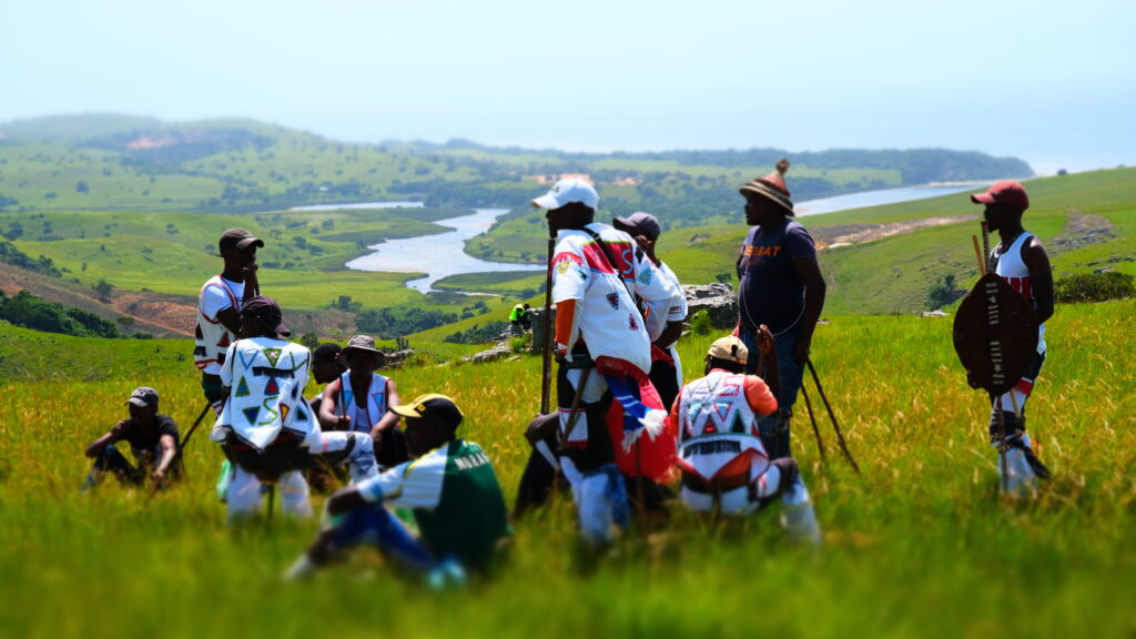 Hombres de la comunidad amaMpondo preparándose para una celebración cultural con la desembocadura de un río y el mosaico paisajístico de praderas y bosques como telón de fondo